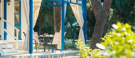 A covered outdoor seating area with a table and chairs, surrounded by trees and plants.