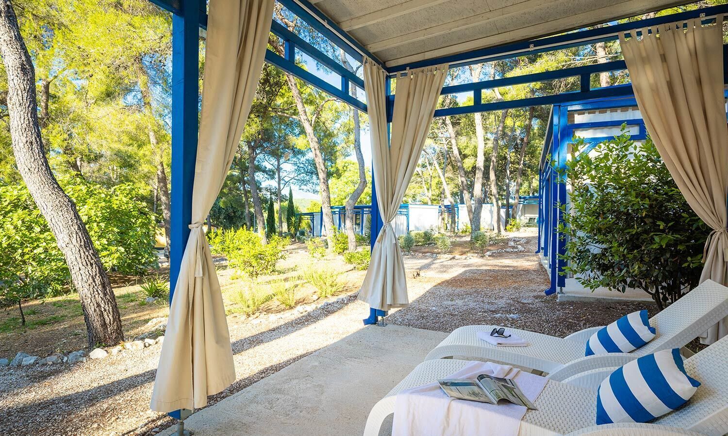 A covered outdoor seating area with beige cushions and striped pillows, surrounded by trees and a gravel path.
