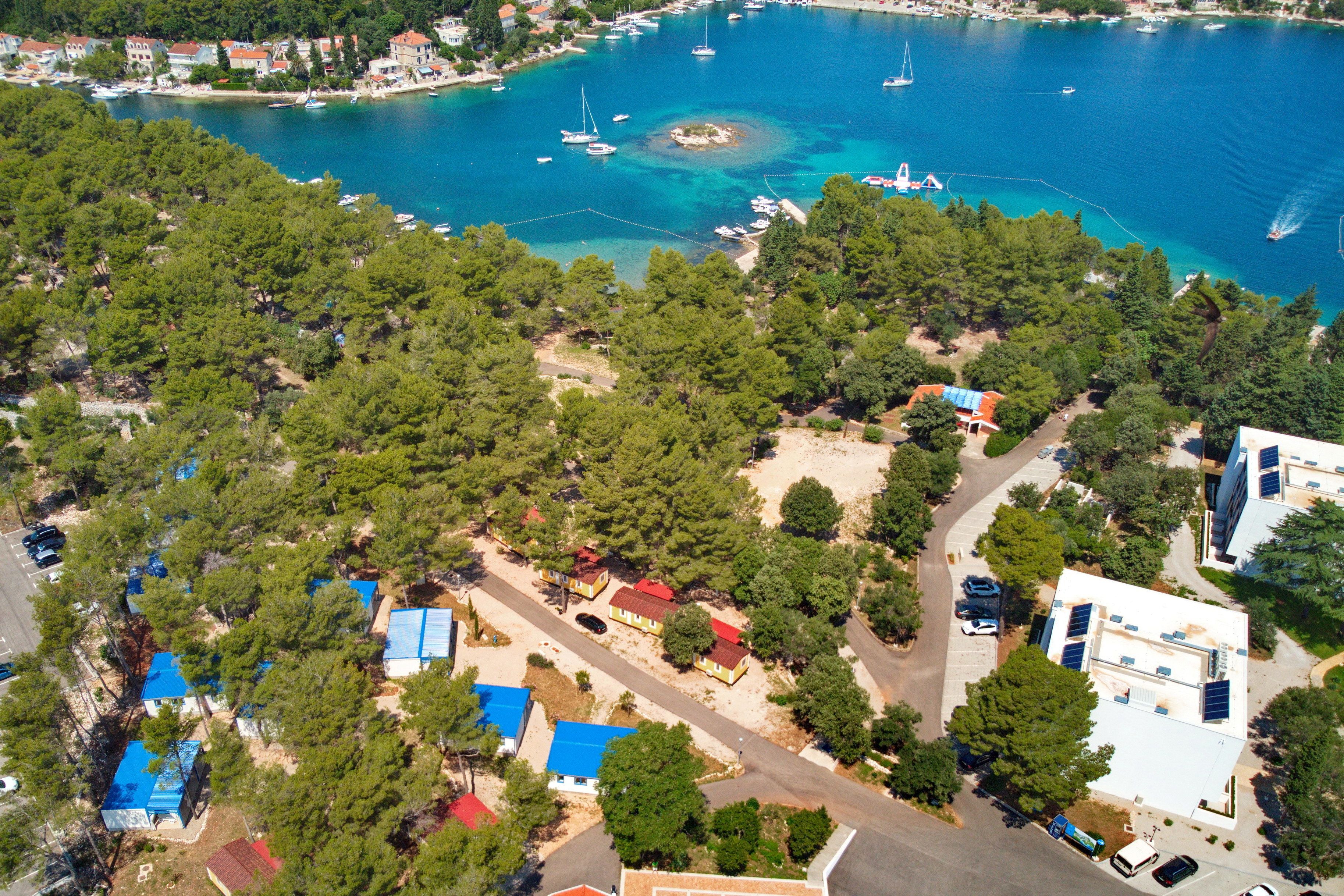 An aerial view of a coastal area with several buildings, some with blue roofs, surrounded by dense green trees and a clear blue sea with boats.