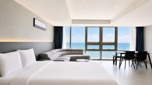 Habitación de hotel moderna con una cama grande, un sofá, una mesa de comedor con sillas y vista al océano.