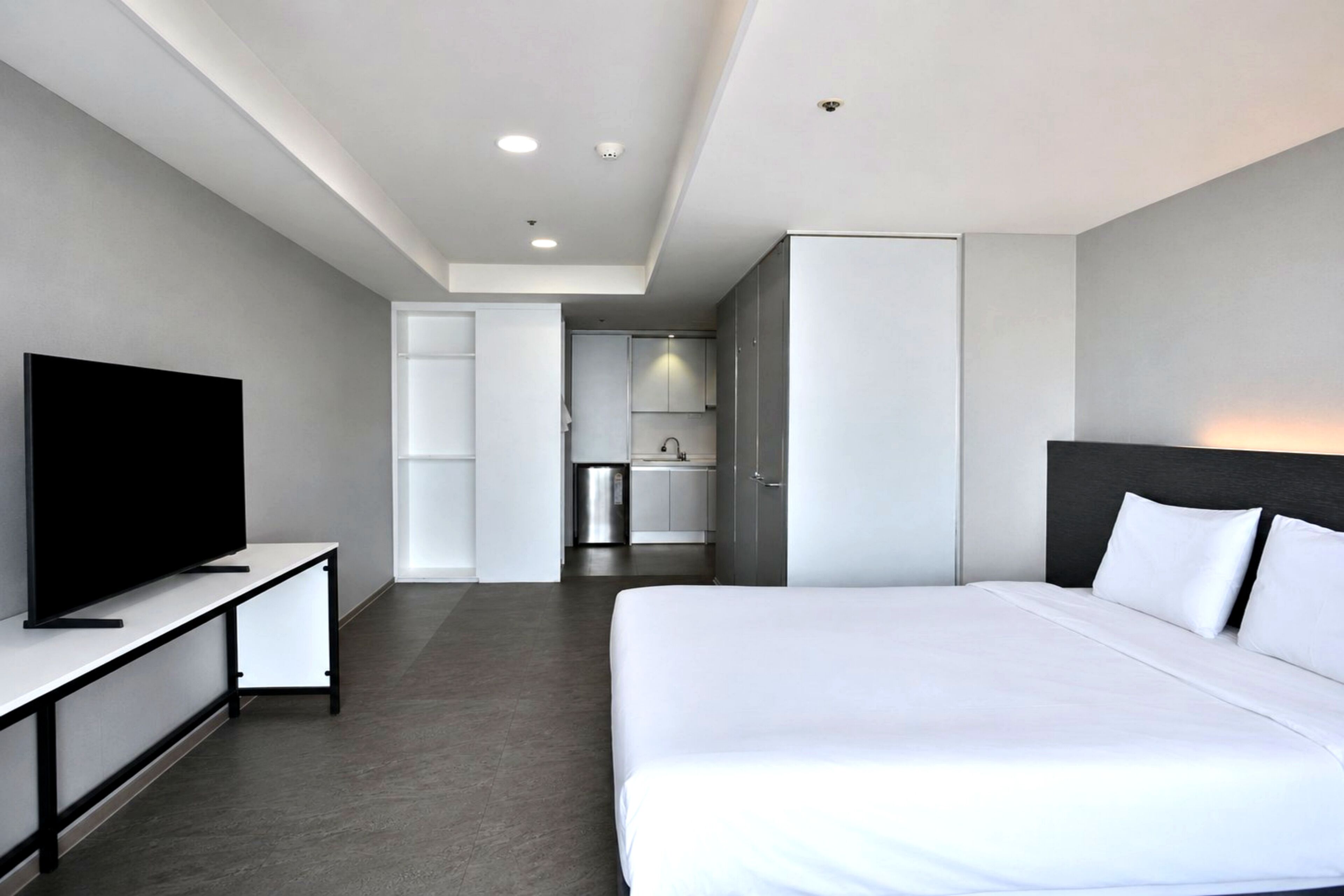 A modern hotel room with a large bed, a TV on a stand, a kitchenette, and a bathroom visible through an open door.