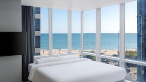 Habitación de hotel con una cama grande, un televisor de pantalla plana y vistas a la playa y al océano a través de ventanales de piso a techo.