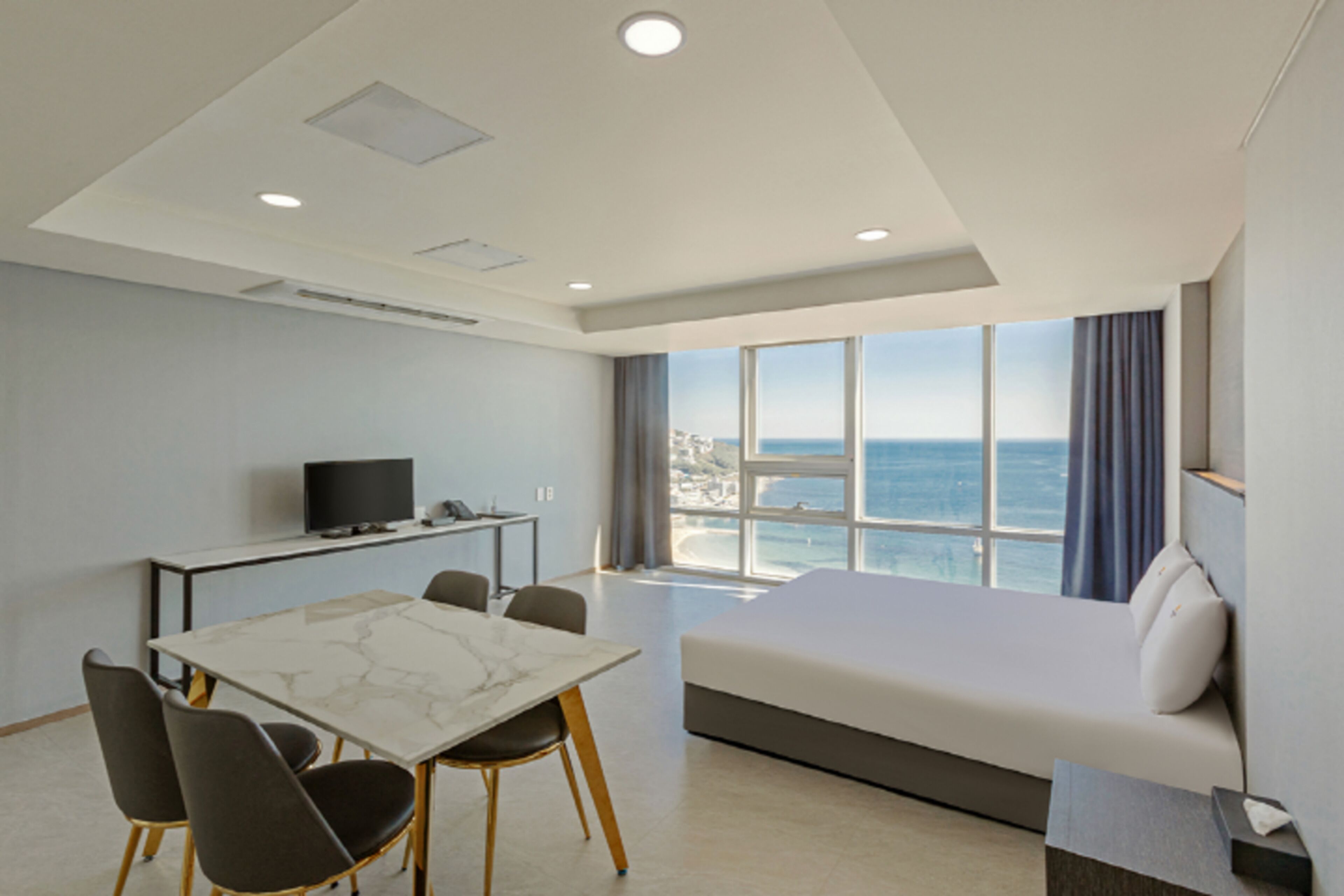 A hotel room with a bed, a desk, a TV, and a view of the sea.