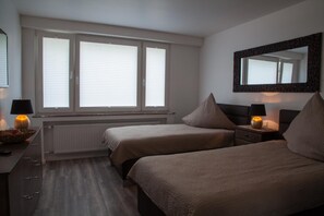 A bedroom with two beds, a dresser, a mirror, and a window with blinds. - Die Hotelalternative (Solingen)