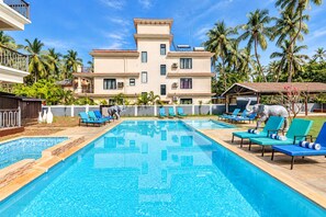 Una piscina con sillas de descanso, palmeras y un edificio de varios pisos al fondo.