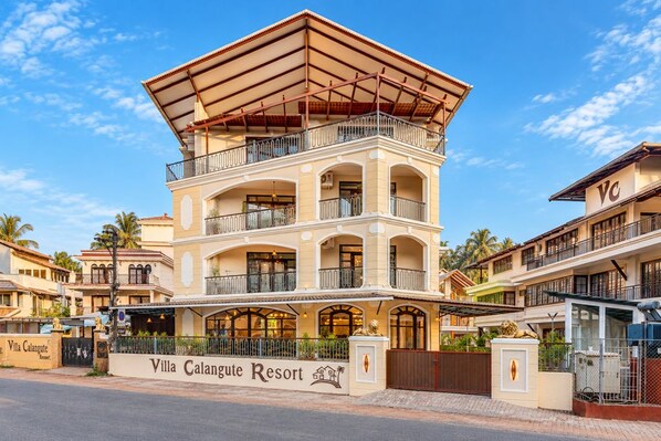 Un hotel de varios pisos con un balcón destacado en el segundo piso, un letrero que dice 'Villa Calangute Resort' y una entrada con rejas.