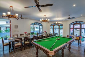 Una sala de billar con mesa de pool, ventiladores de techo, candelabros y amplias ventanas.