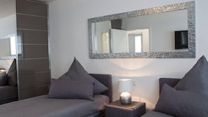 A modern bedroom with a large mirror, two beds with grey bedding, a bedside table with a lamp, and a television on the wall. - Luxusappartement - Die Hotelalternative (Solingen)