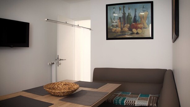 A modern room with a wooden dining table, a woven basket, a brown sofa with patterned cushions, a flat-screen TV, and a framed painting of bottles and pumpkins on the wall. - Luxusappartement - Die Hotelalternative (Solingen)