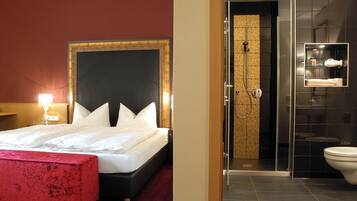 Habitación de hotel con una cama grande, un banco rojo, zona de ducha y baño con cabina de vidrio.