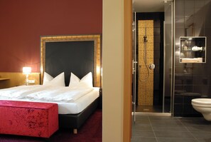 Une chambre d’hôtel avec un grand lit, un banc rouge, un espace douche et une salle de bain dotée d’une paroi en verre.