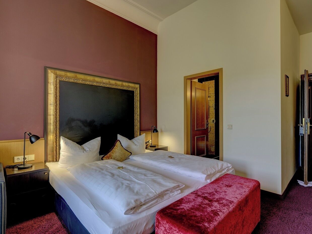 Habitación de hotel con una cama grande, una mesita de noche, un banco rojo y una puerta que da a otra habitación.