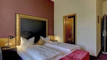 Habitación de hotel con una cama grande, una mesita de noche, un banco rojo y una puerta que da a otra habitación.