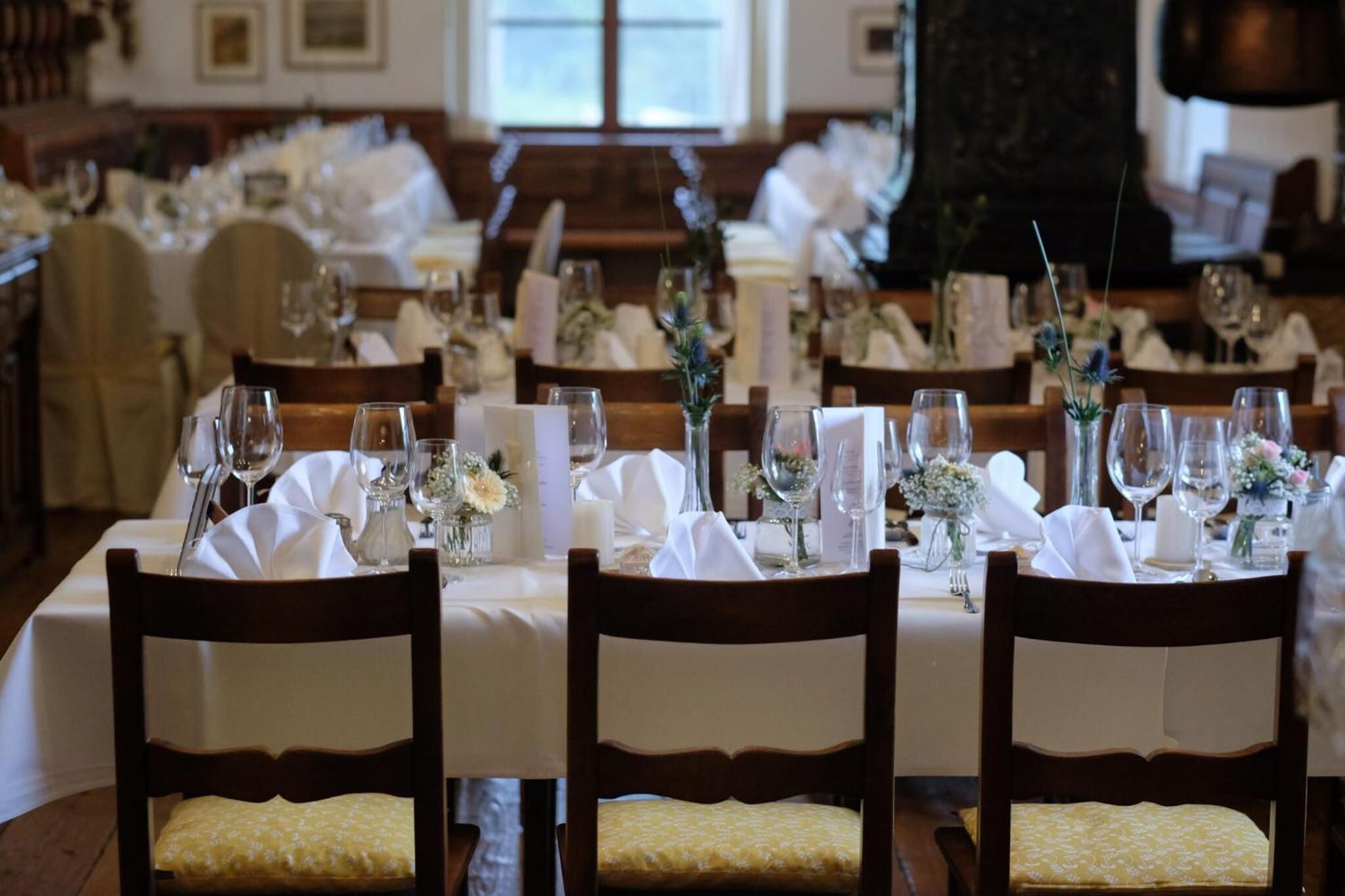 A formal dining room with multiple tables set for a meal, each with white tablecloths, glassware, and floral centerpieces.