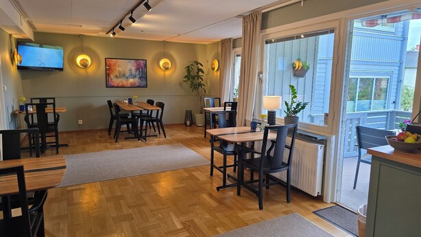 A modern dining area with wooden tables and chairs, a flat-screen TV mounted on the wall, and a large window with curtains. - Hotell Zlafen B&B (Karlskoga)