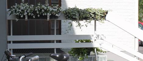 Un edificio blanco con puerta de madera, ventana con reja metálica y balcón con macetas de flores.