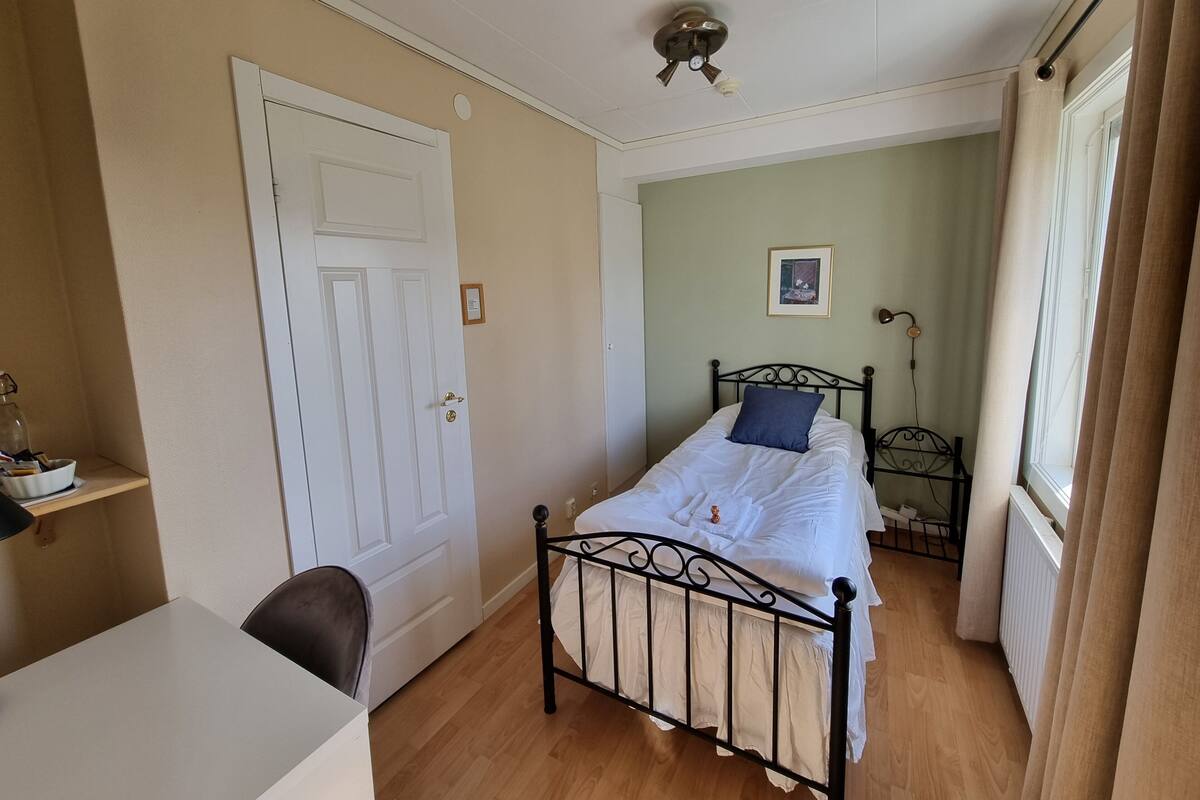 A bedroom with a bed, a desk, a chair, a radiator, and a window with curtains.