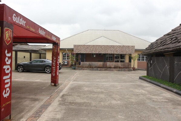 A parking area with a car, a building with a tiled roof, and a red banner with the text "Gulder". - Kastrufid Lodge (Uyo)