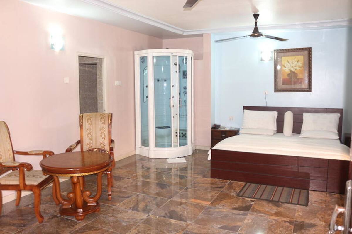 Habitación de hotel con cama, ventilador de techo, ducha con mampara de vidrio, mesa de comedor con sillas y un cuadro en la pared.