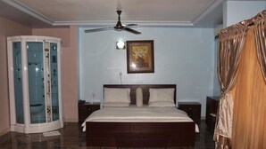 Une chambre avec un grand lit, un ventilateur de plafond, une cabine de douche en verre et un tableau encadré au mur.