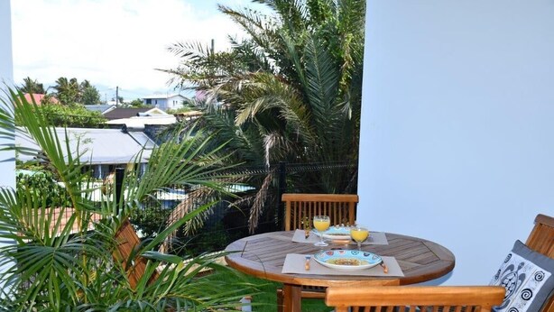 Une table et des chaises en bois sont disposées à l’extérieur, offrant une vue sur un quartier résidentiel et des palmiers.