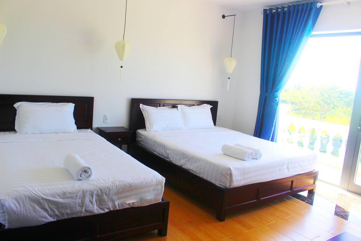 A hotel room with two beds, wooden headboards, white linens, and a view of greenery through a large window.