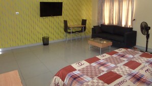 A room with a yellow patterned wall, a flat-screen TV mounted above a dining table with chairs, a black sofa, a glass coffee table, and a bed with a checkered bedspread. - Megsplace Inn (Lagos)