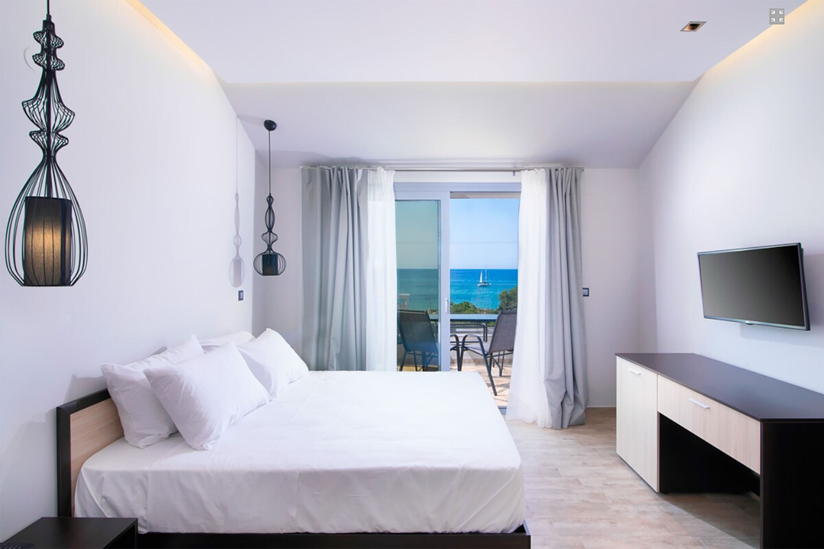 Een hotelkamer met een bed, een bureau, een televisietoestel en een balkon met uitzicht op zee.