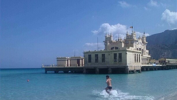 A beach with clear turquoise water, a pier, and a large building with multiple towers. - Holiday House Partanna Mondello (Mondello)