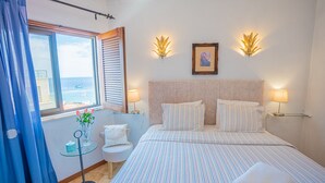 A hotel room with a bed, bedside lamps, a small table with a vase of flowers, a window with a view of the sea, and a picture on the wall.