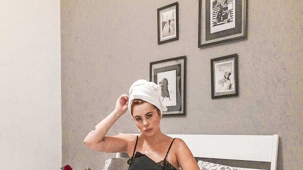 Una persona sentada en una cama con un libro, una taza y una toalla en la cabeza. En la pared hay fotografías enmarcadas y una mesita con un cuenco de comida.