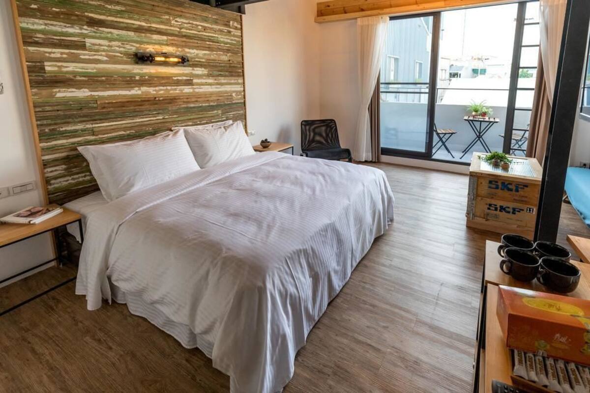 A bedroom with a large bed, a wooden headboard, a small table with a plant, and a view of the outside through a large window.