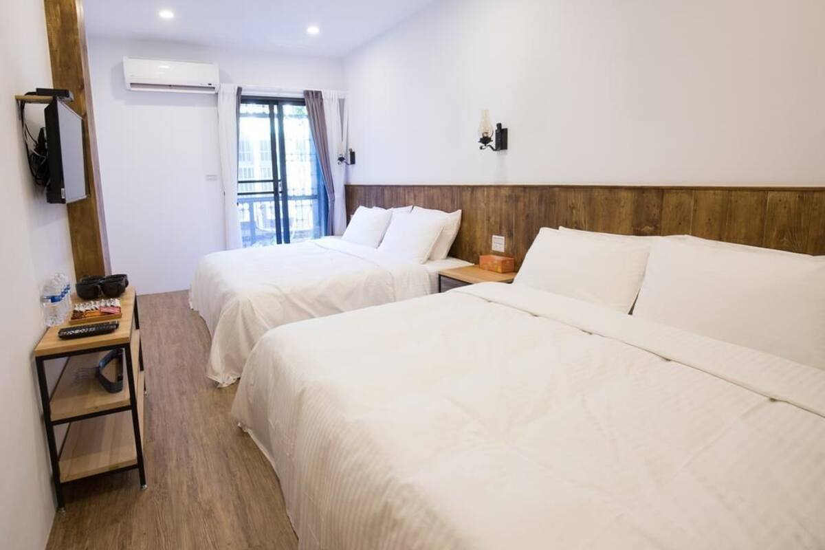 A hotel room with two single beds, a wooden headboard, a small table with a lamp, and a TV mounted on the wall.
