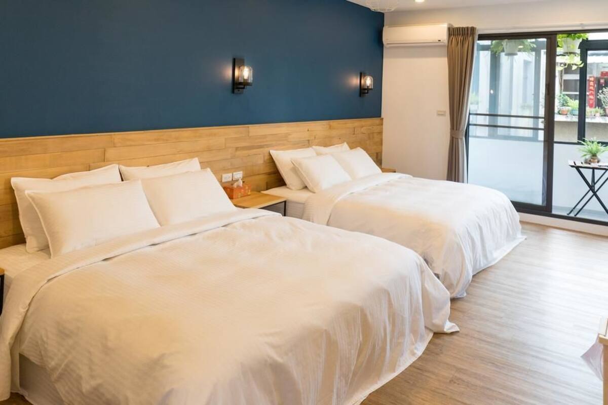 A hotel room with two beds, a wooden headboard, a blue accent wall, and a view of the outside through a large window.