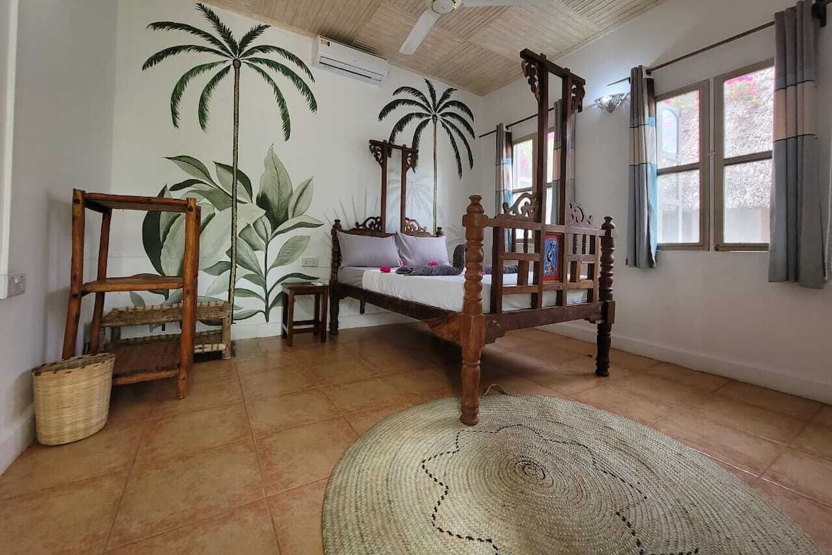 Une chambre avec un lit en bois, un ventilateur de plafond, un grand tapis tissé et un mur décoré de peintures murales représentant des palmiers.