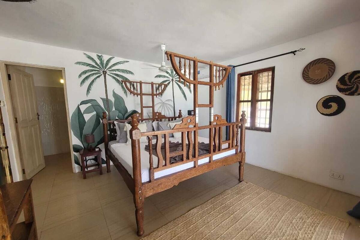 Une chambre à coucher avec un lit en bois, une petite table, une chaise, une fresque murale et une fenêtre avec des rideaux.