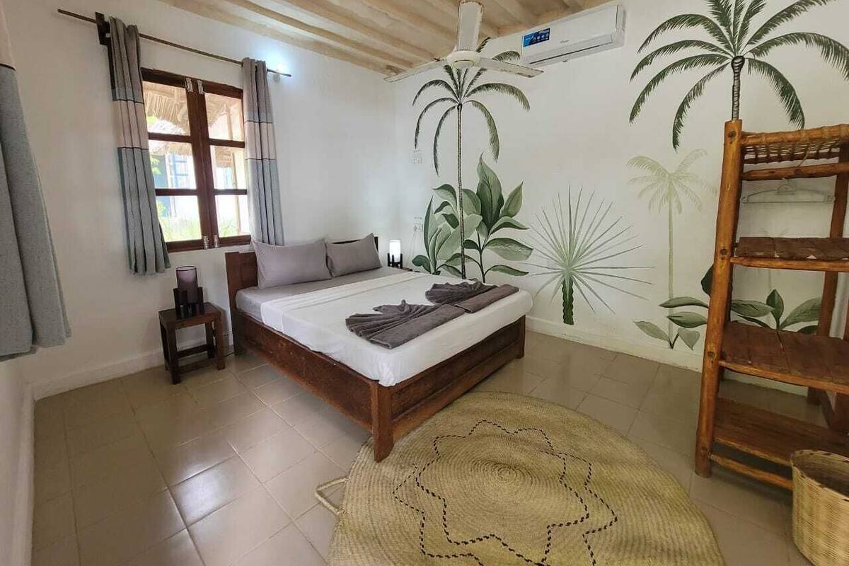 Une chambre à coucher avec un lit, un ventilateur de plafond, une fenêtre avec des rideaux et une fresque murale représentant des palmiers.