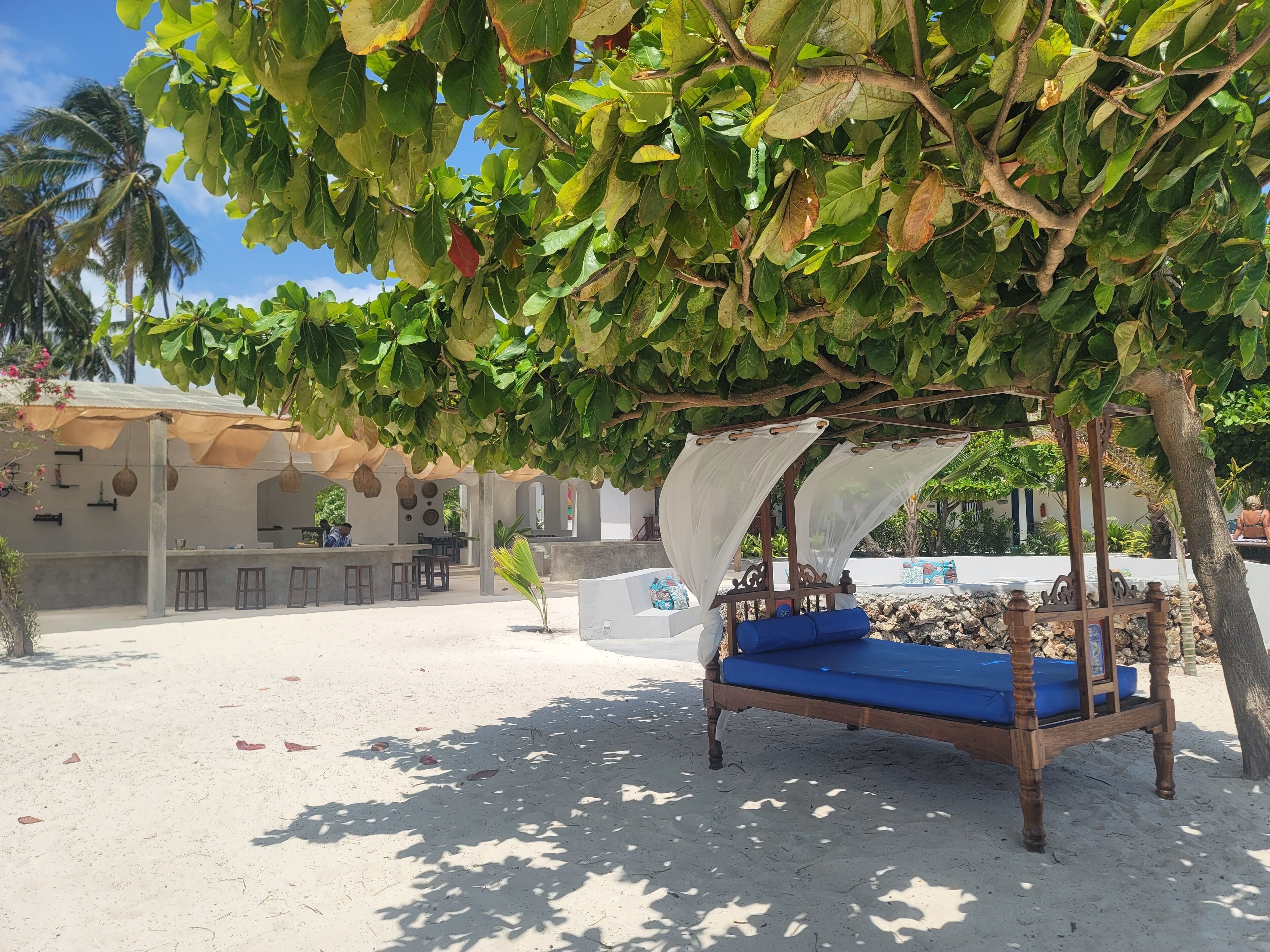 A beachside outdoor seating area with a wooden canopy bed, a bar, and a covered seating area with tables and chairs.
