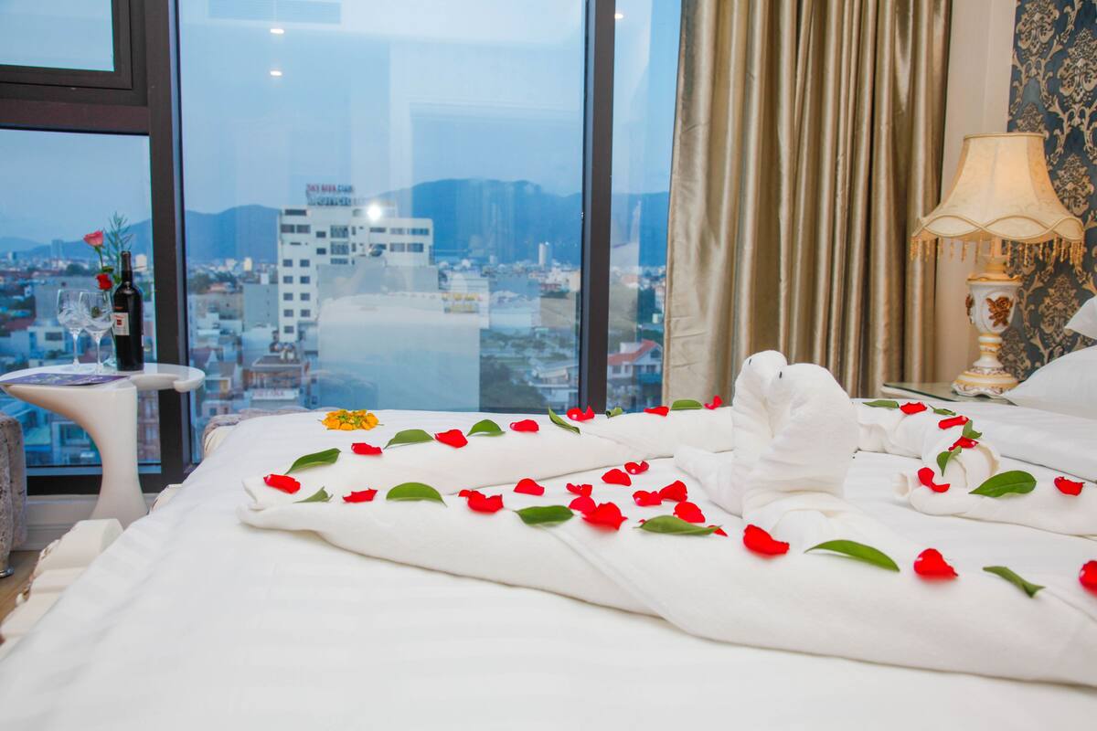 Habitación de hotel con una cama decorada con pétalos de rosa, vistas a una ciudad a través de amplias ventanas y una mesita de noche con una lámpara.