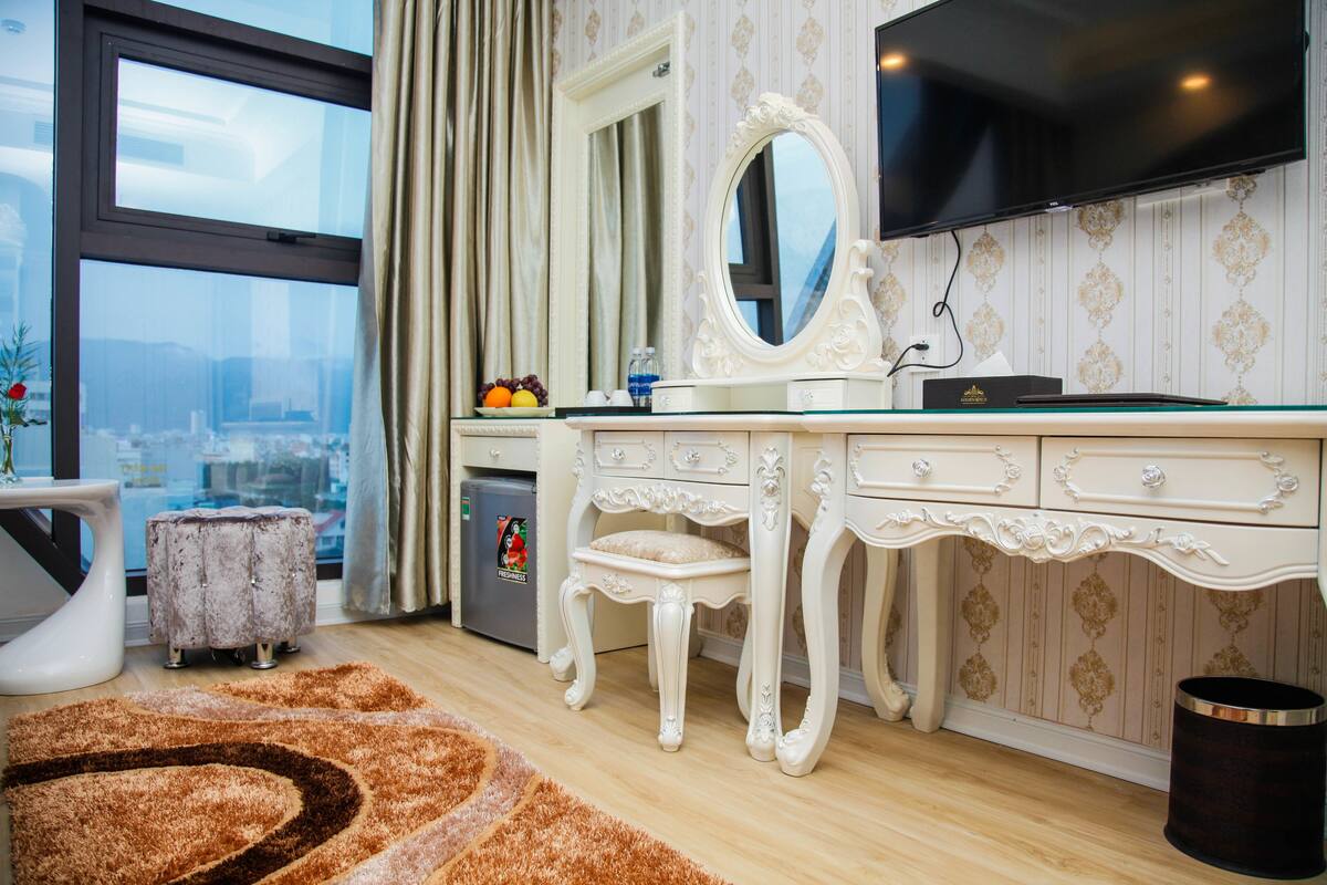 Una habitación con un ventanal grande, un tocador con espejo, un taburete pequeño, un cesto de basura y una alfombra decorativa.