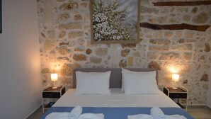 Une chambre d’hôtel avec un mur en pierre, un lit recouvert de draps blancs, deux tables de chevet avec des lampes et un tableau représentant des fleurs accroché au mur.