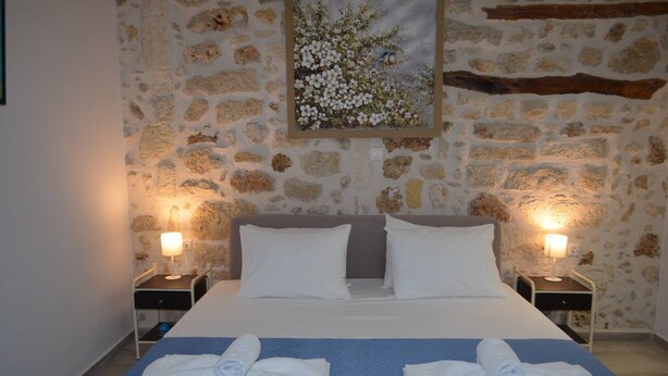 Une chambre d’hôtel avec un mur en pierre, un lit recouvert de draps blancs, deux tables de chevet avec des lampes et un tableau représentant des fleurs accroché au mur.