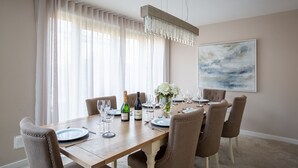 Une salle à manger avec une table en bois dressée pour six personnes, agrémentée d’un lustre, d’un grand tableau abstrait et d’une vue sur l’extérieur à travers des rideaux légers.