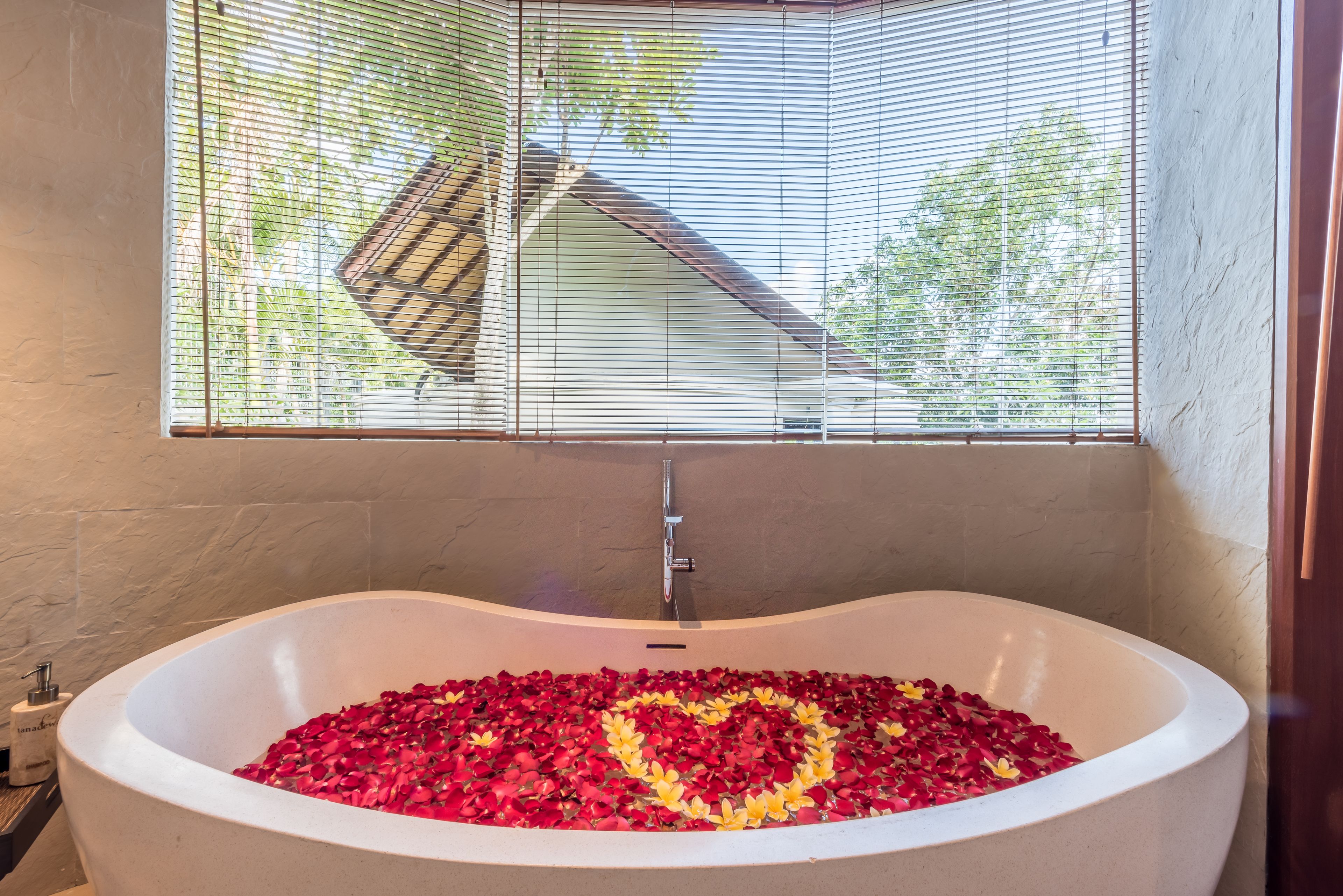 A bathtub filled with rose petals arranged in a heart shape, a window with blinds, and a bathroom with a towel dispenser.