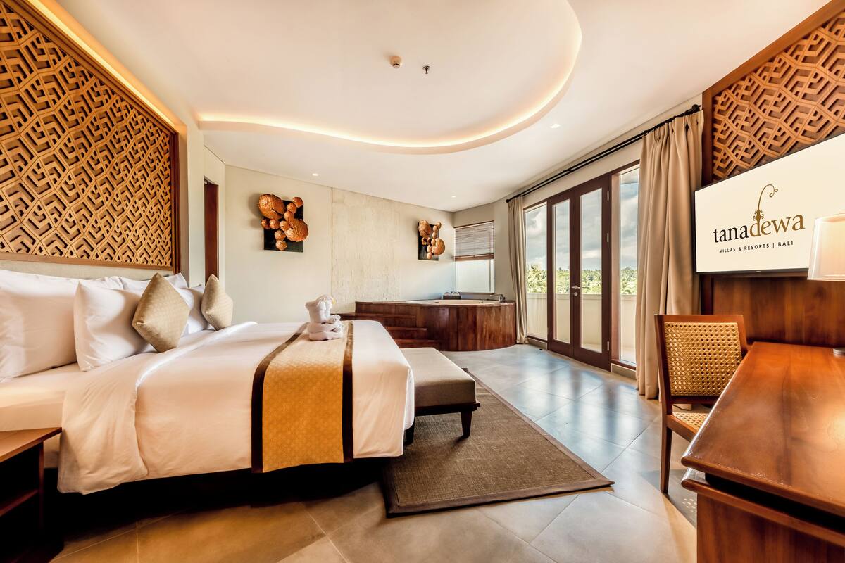 A modern hotel room with a large bed, a wooden headboard, a dining area, and a view of the outdoors.