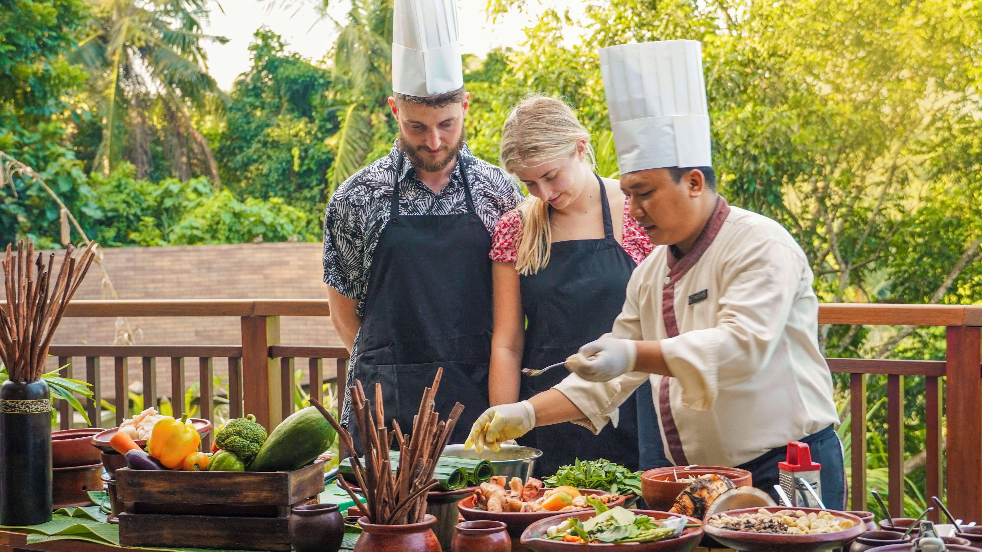 A chef preparing food outdoors with a variety of vegetables and spices on a wooden table.