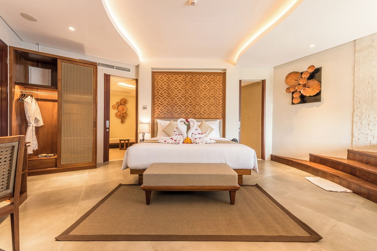 A modern hotel room with a large bed, a wooden bench, a walk-in closet, and a decorative wall piece.