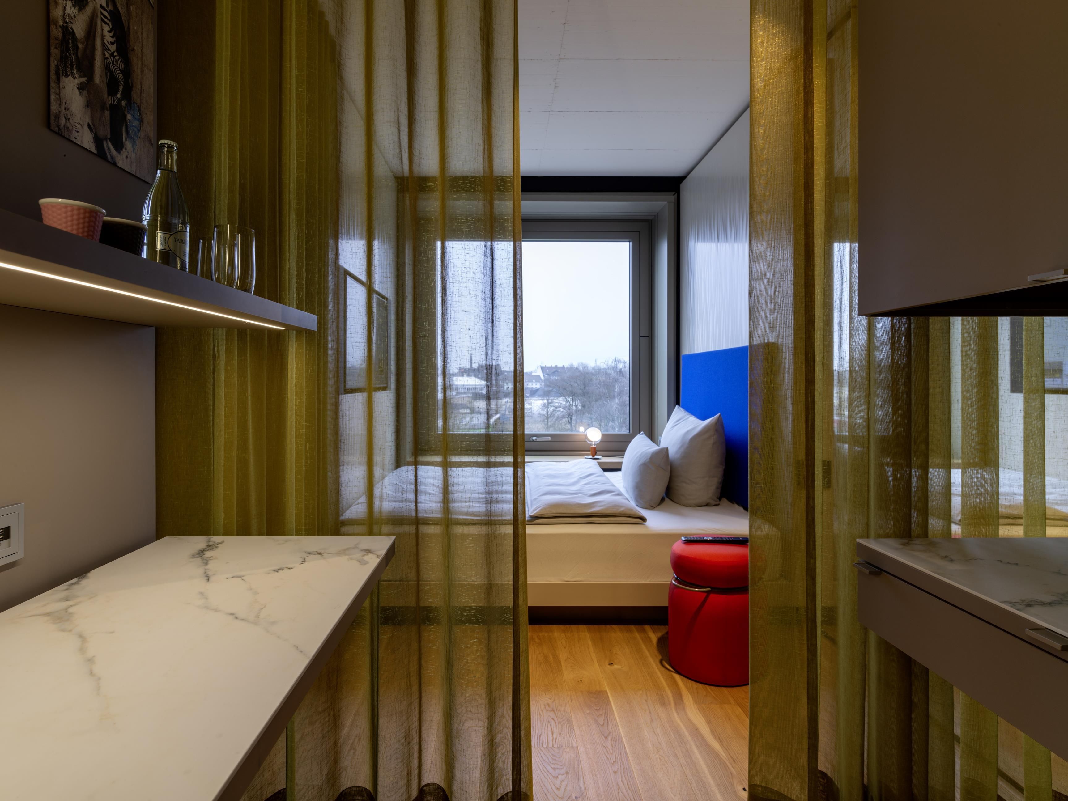 Un dormitorio moderno con una cama, un reposapiés rojo y una ventana con vistas a un paisaje urbano.