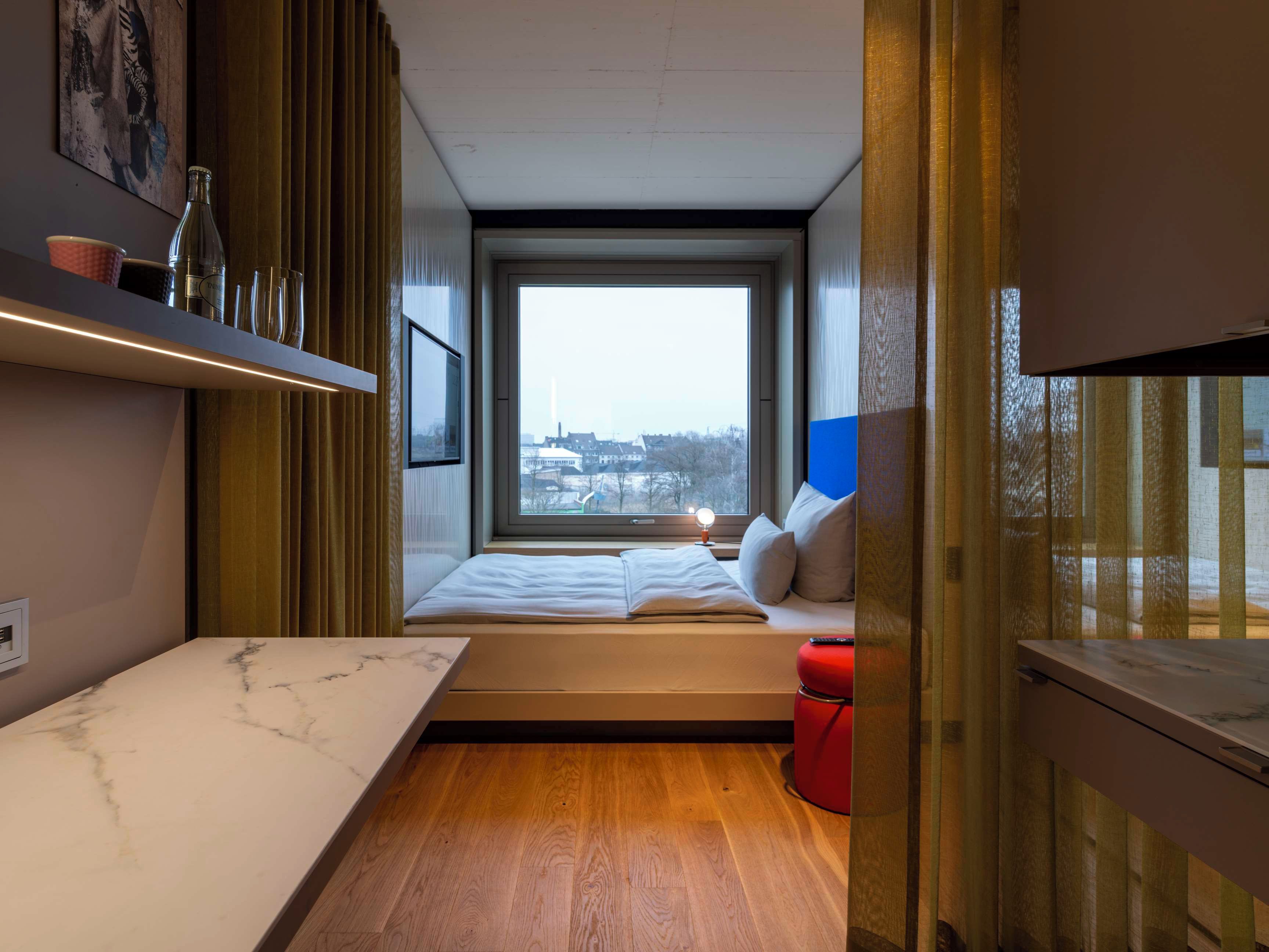 Una habitación pequeña con cama, una mesita y ventana con vistas a la ciudad.