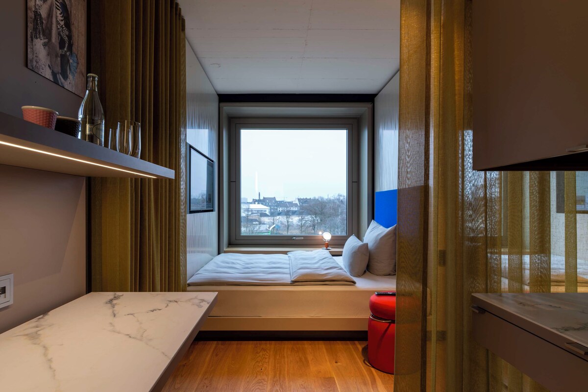 Una stanza compatta con un letto, un tavolino e una finestra vista città.